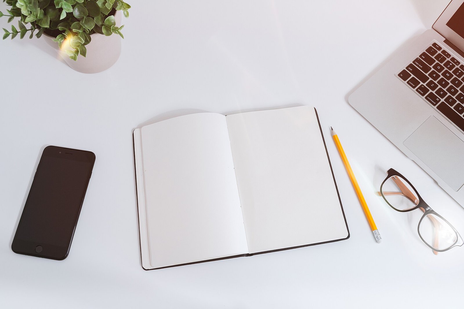 Open blank notebook, yellow pencil, glasses, smartphone and laptop on a white desk